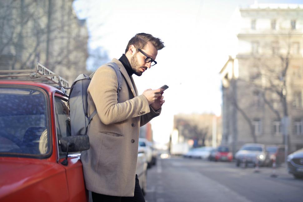 Man in Beige Coat Holding Phone Leaning on Red Vehicle