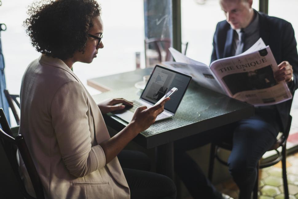 Free Image of Two businesspeople at a cafe, looking at mobile phone and newspaper 
