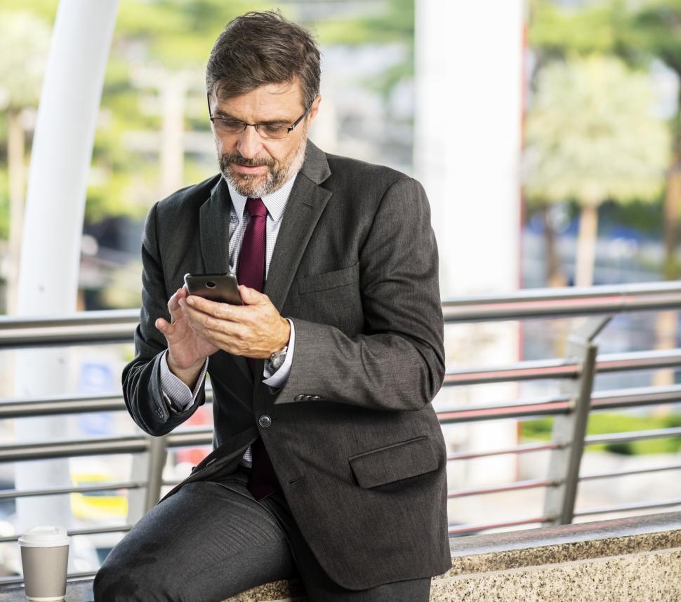 Free Image of A bearded businessman operating his mobile phone sitting on a railing 