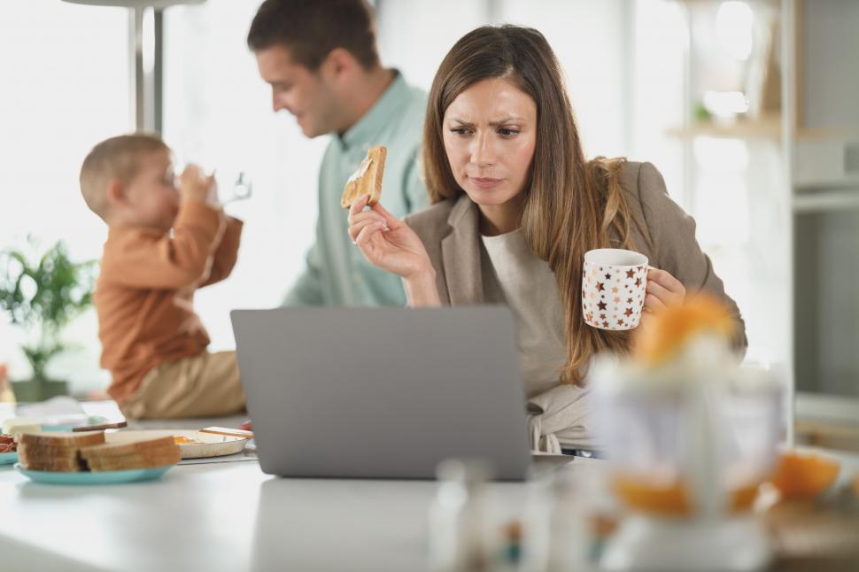 Free Image of Young mother holding coffee cup and eating toast while using laptop 