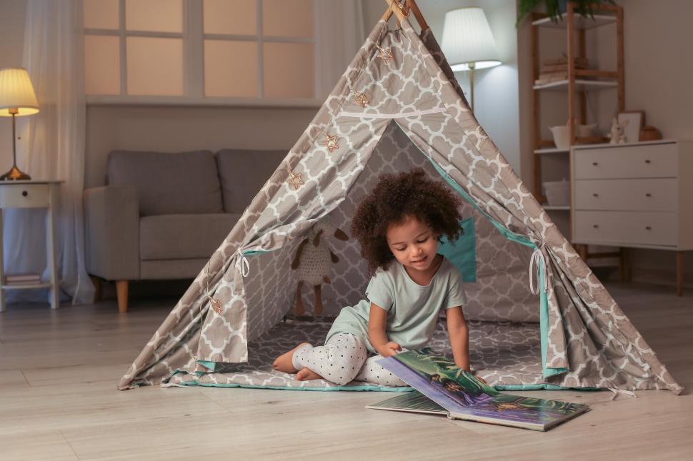 Little daughter sitting with book in a camping tent at home