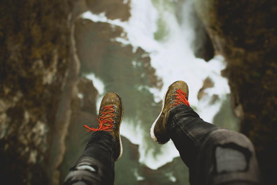 Person wearing hiking boots standing above a beautiful waterfall surrounded by lush forest and mist