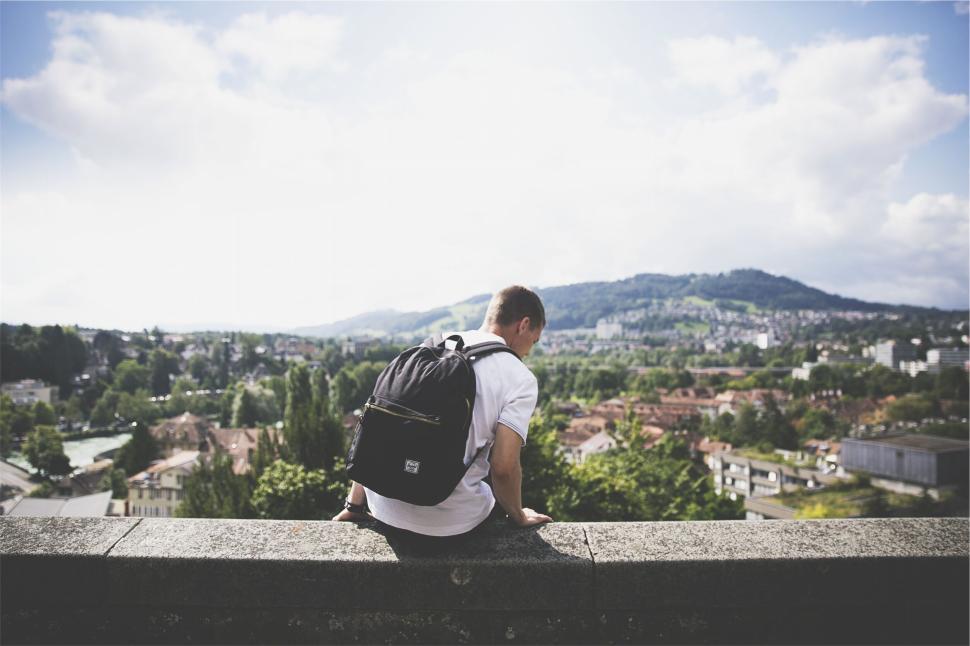 Free Image of Man sitting with a backpack overlooking a scenic town 
