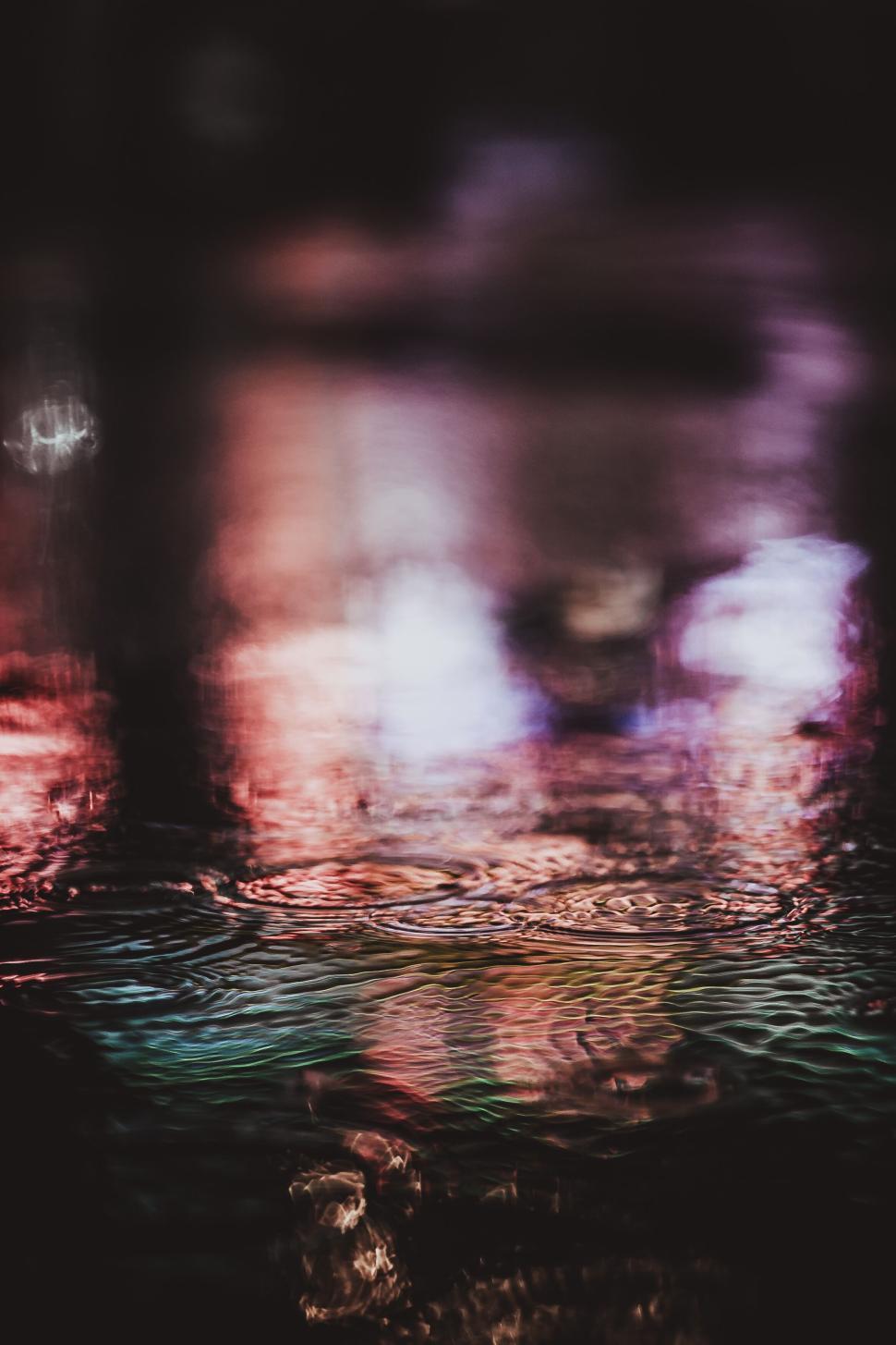 Free Image of Raindrops creating ripples on a puddle reflecting city lights. 
