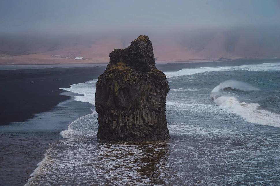 Free Image of Rock formation on a black sand beach with waves crashing around 
