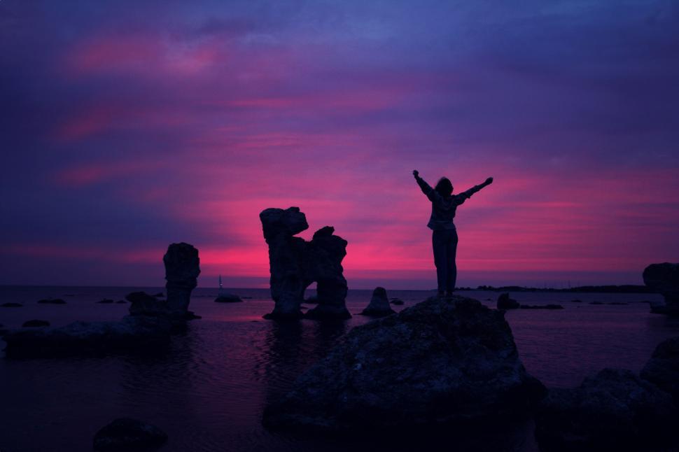 Free Image of Silhouette of a person with raised arms on rocks at purple sunset. 