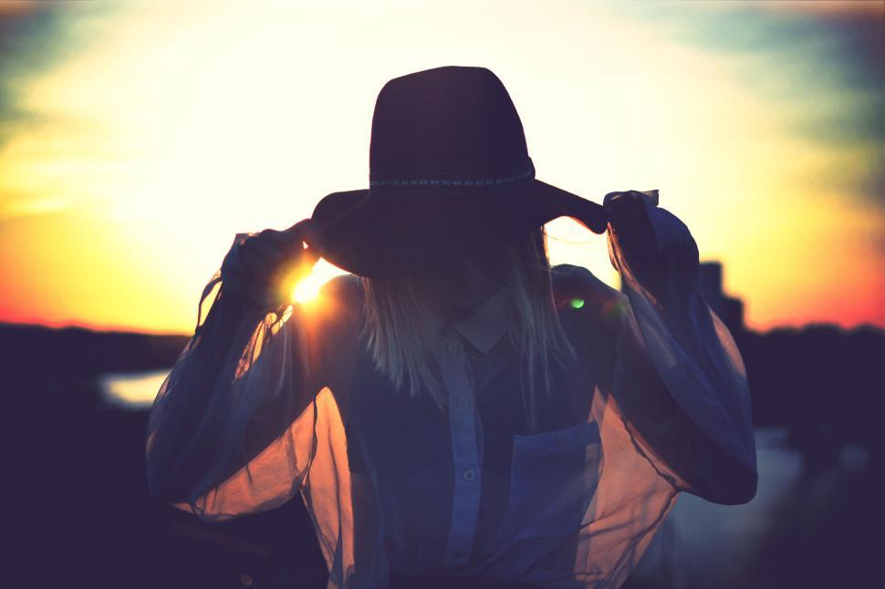Free Image of Woman in hat adjusting her hair with sun setting in the background 