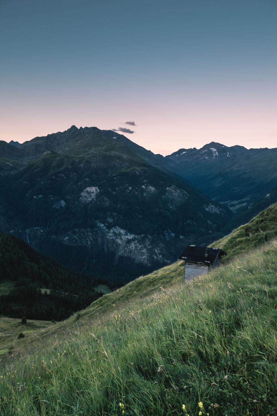 Free Image of Small cabin on a grassy hill with mountain range in background. 