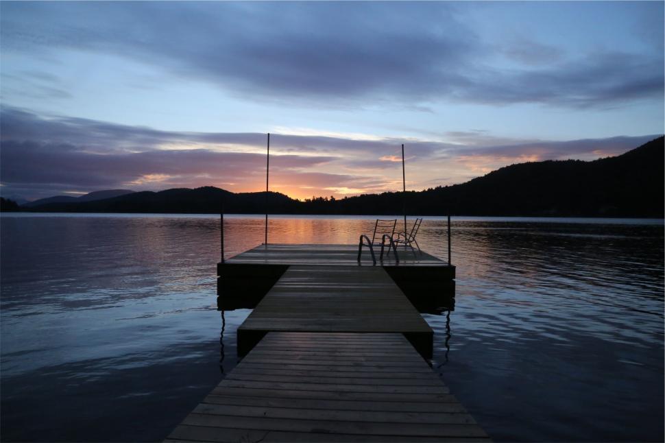 Free Image of Dock extending into calm lake at sunset with mountains silhouette 
