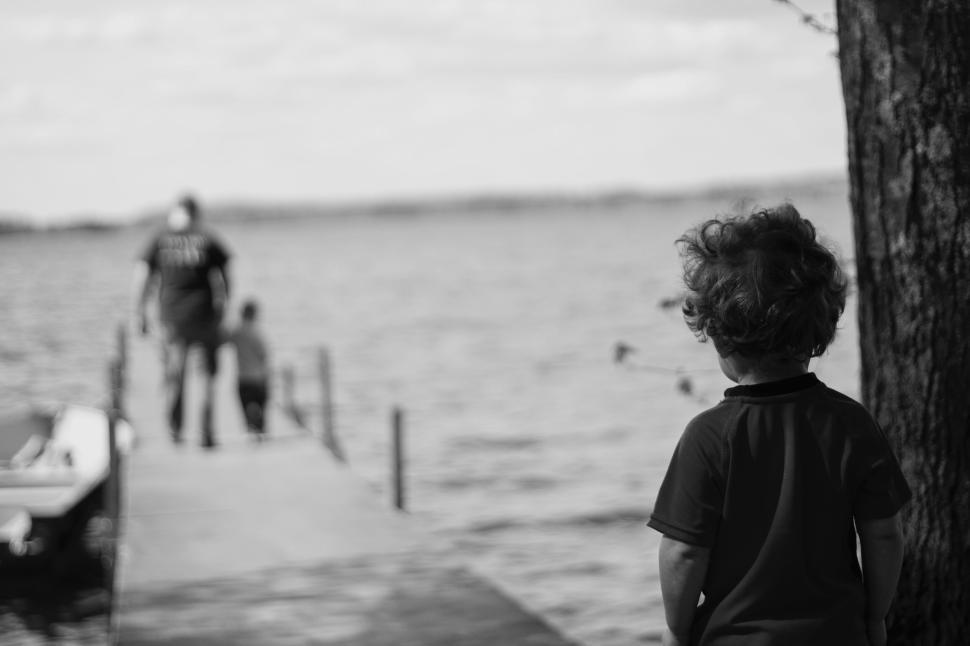 Free Image of Boy looking at people walking on a pier by a lake in black and white 