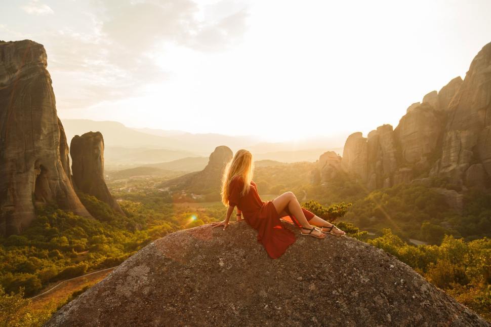 Free Image of Woman in red dress sits on rock with stunning cliff view at sunset. 