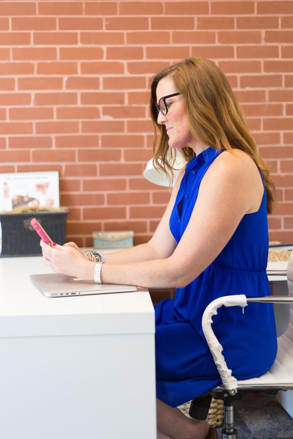 Free Image of Woman in blue dress sitting at desk checking her mobile phone. 