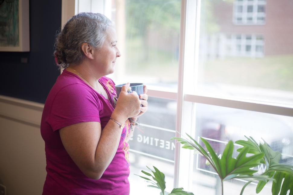 Free Image of Woman holding a cup of coffee looks out of window next to a plant. 