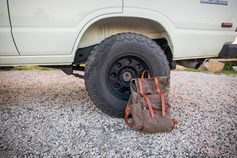 Free Image of Backpack resting by the wheel of a white vehicle on a gravel road. 