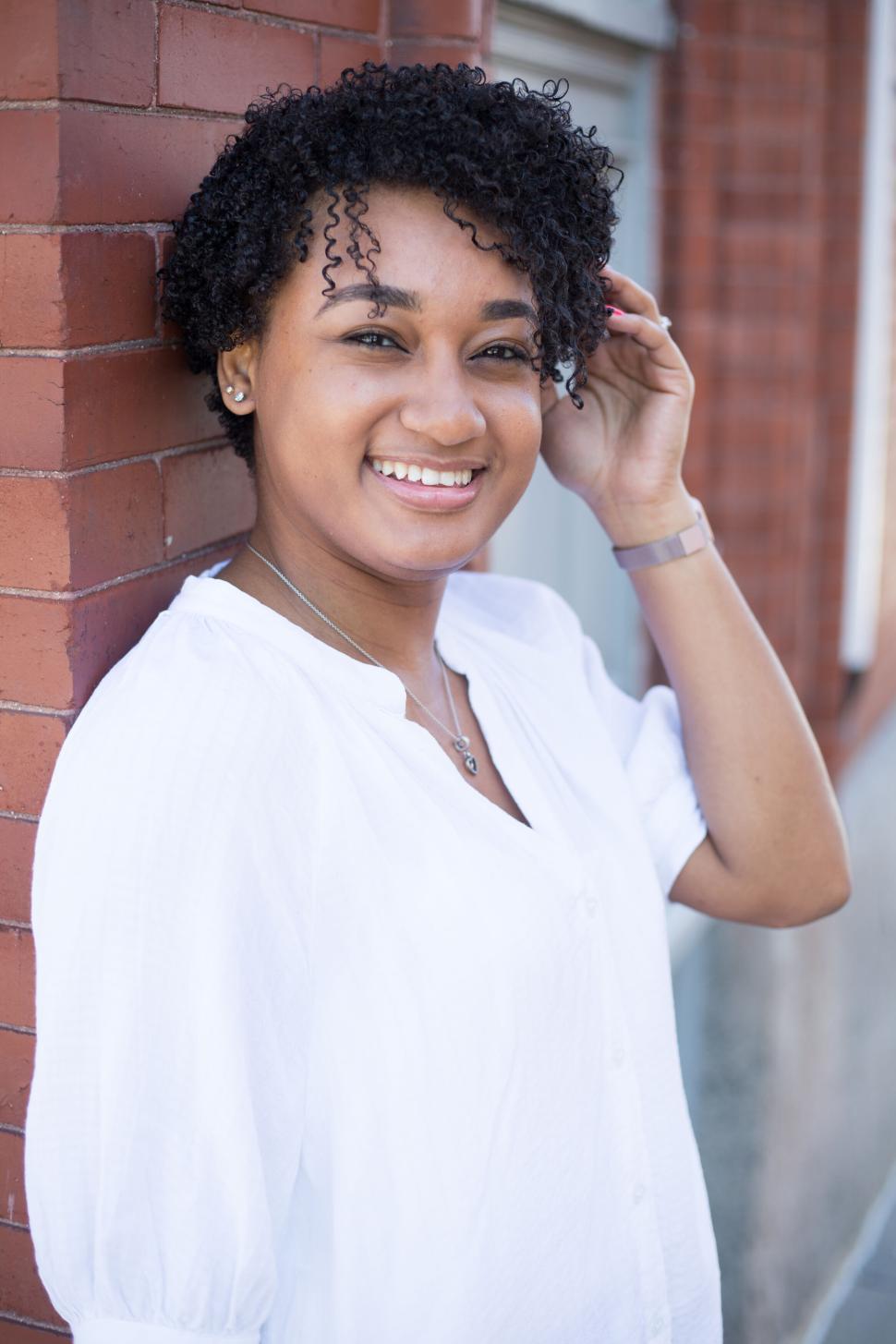 Free Image of Smiling woman posing against brick wall wearing casual outfit. 