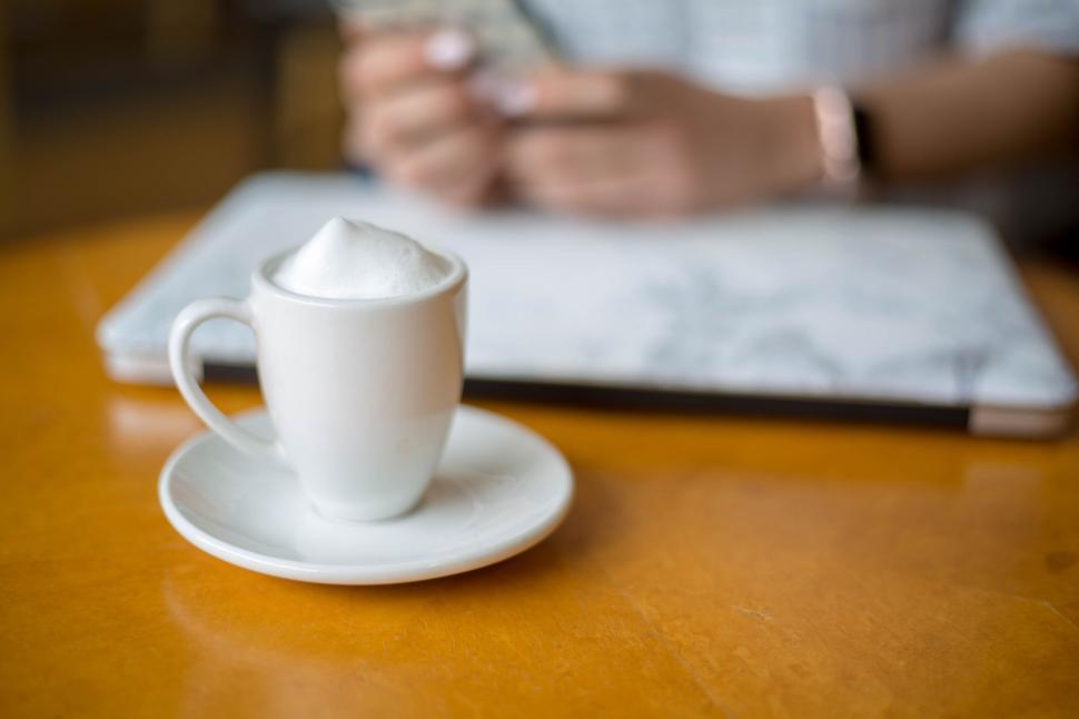 Free Image of Cappuccino cup on table with person using phone and laptop in background 