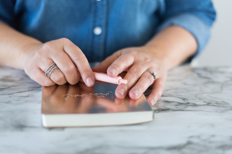 Free Image of Person holding pink pen over bronze notebook on marble surface. 