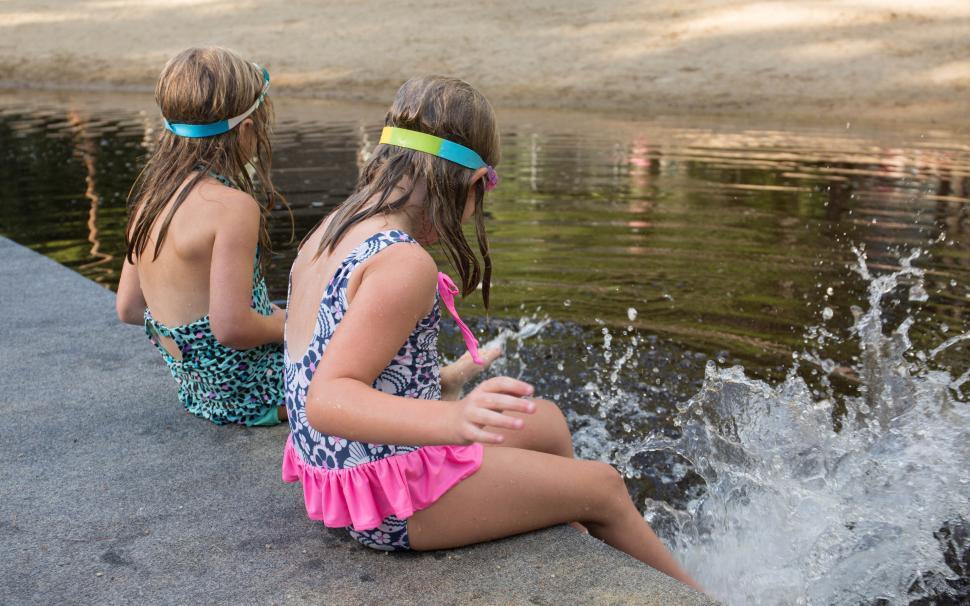 Free Image of Two kids in swimsuits splashing water while sitting by a lake 
