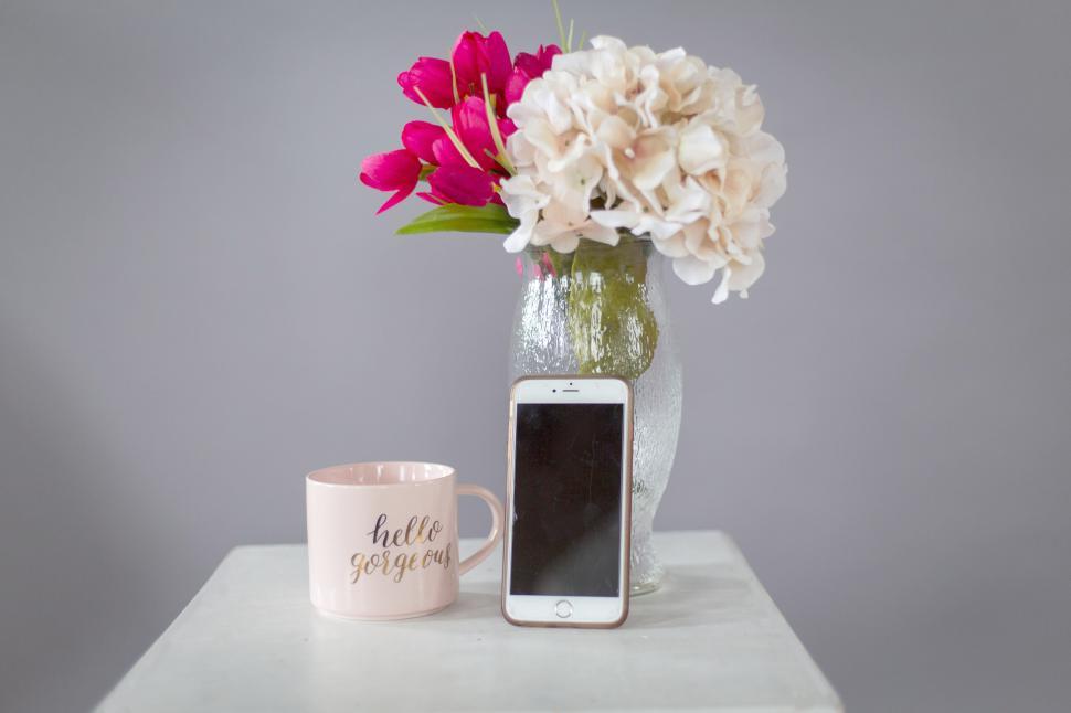 Free Image of Pink mug with  hello gorgeous , white phone, and flowers in vase. 