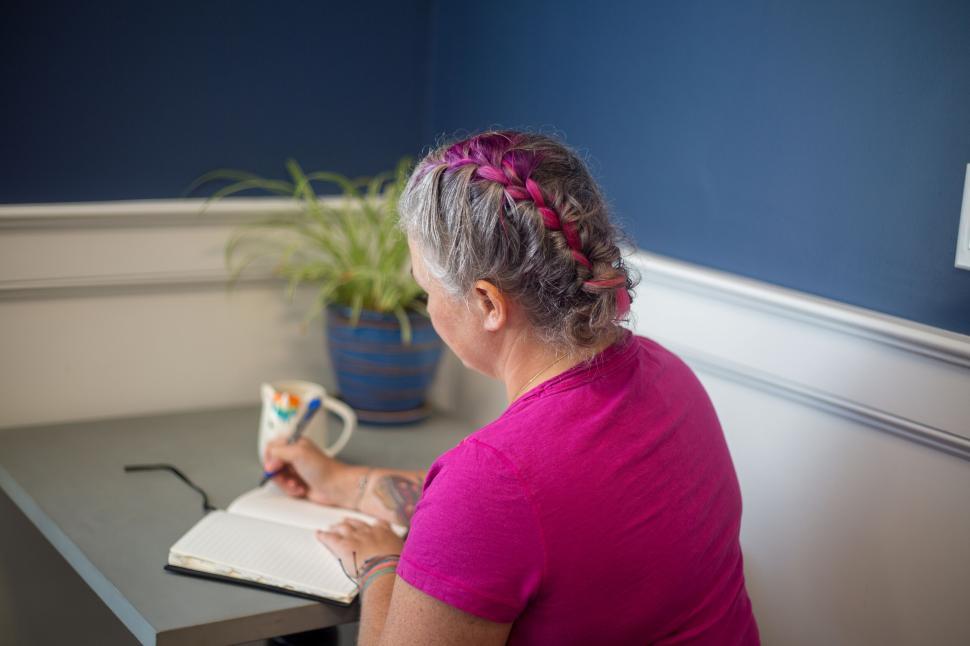 Free Image of Woman with purple braid writing in a notebook at a desk 