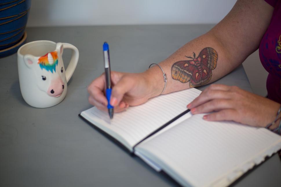 Free Image of Woman writing in journal with a butterfly tattoo and unicorn mug. 