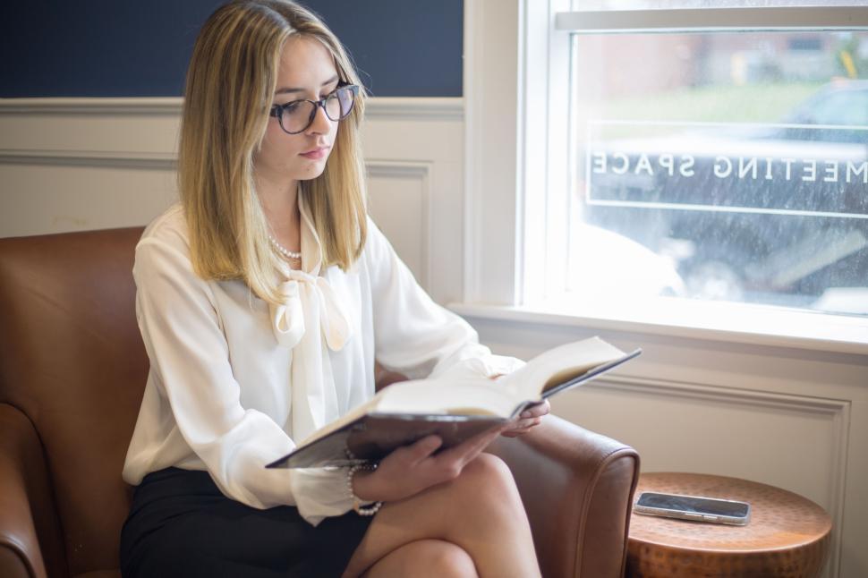 Free Image of Woman reading a book in an elegant office setting on brown chair. 