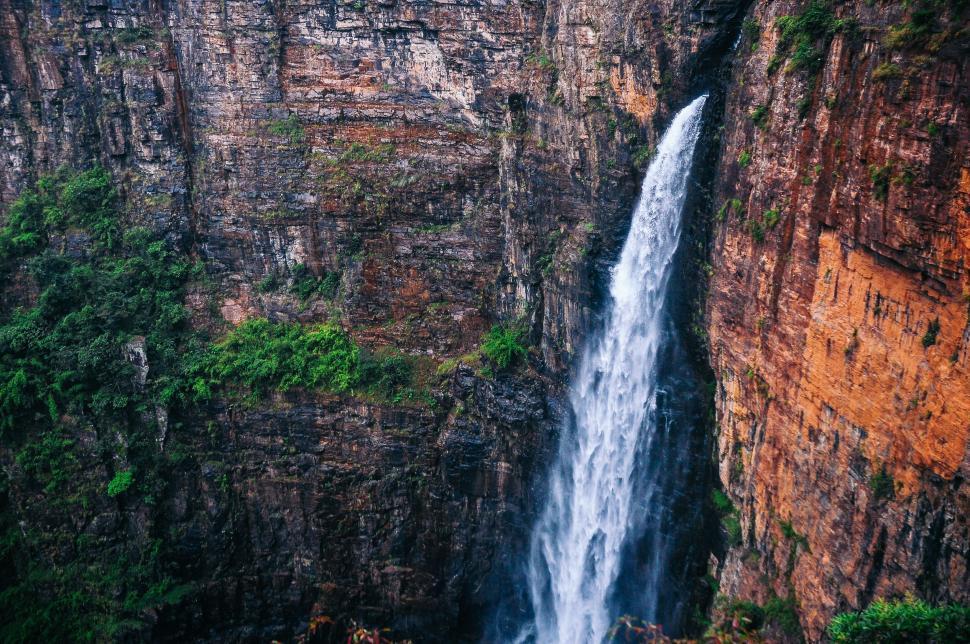 Free Image of Tall waterfall cascading down a rugged cliff surrounded by greenery. 