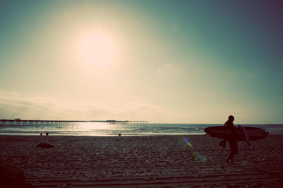 Free Image of Surfer carrying surfboard on scenic beach at sunset with pier 
