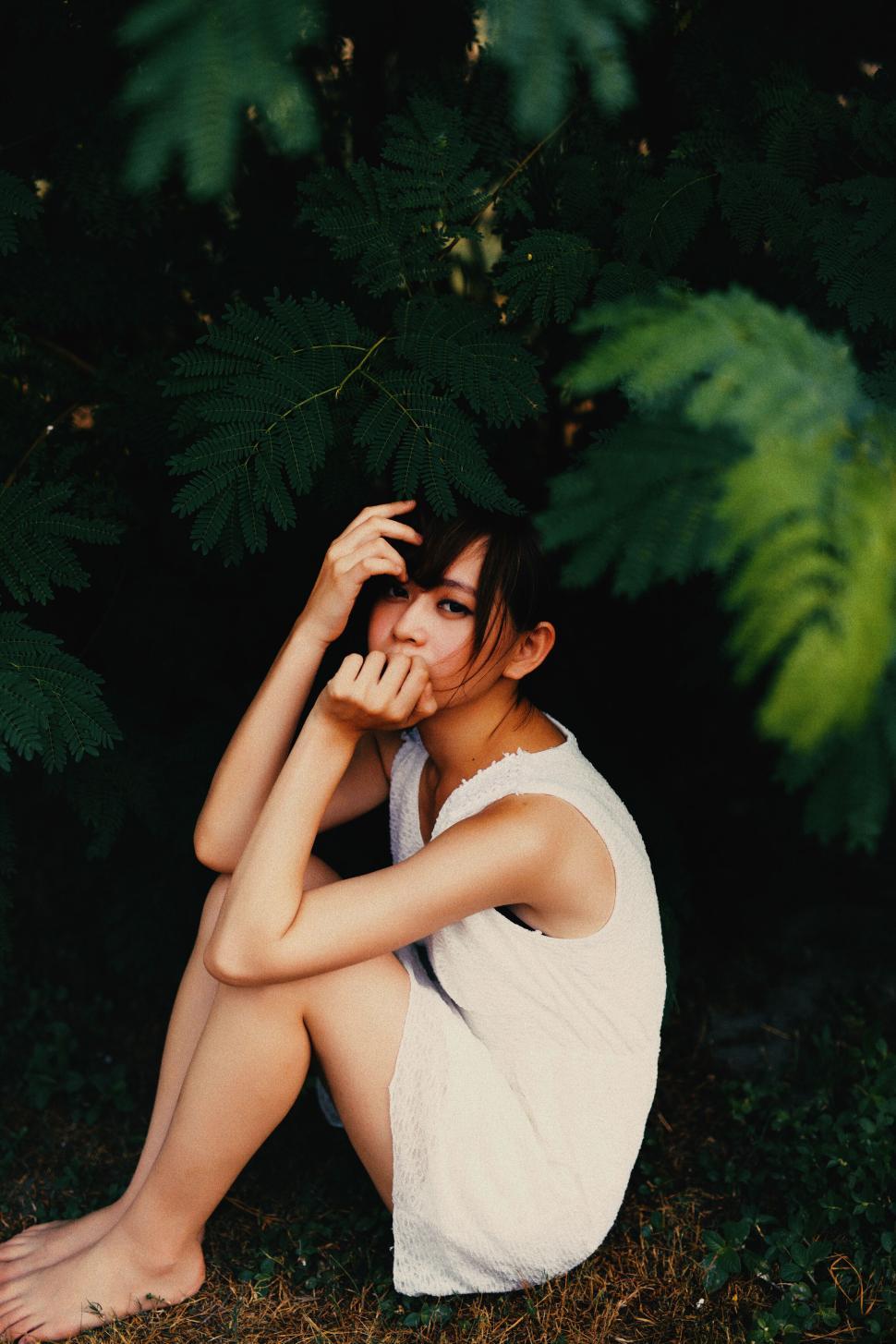 Free Image of Person in white dress sitting on grass under shade of green tree. 