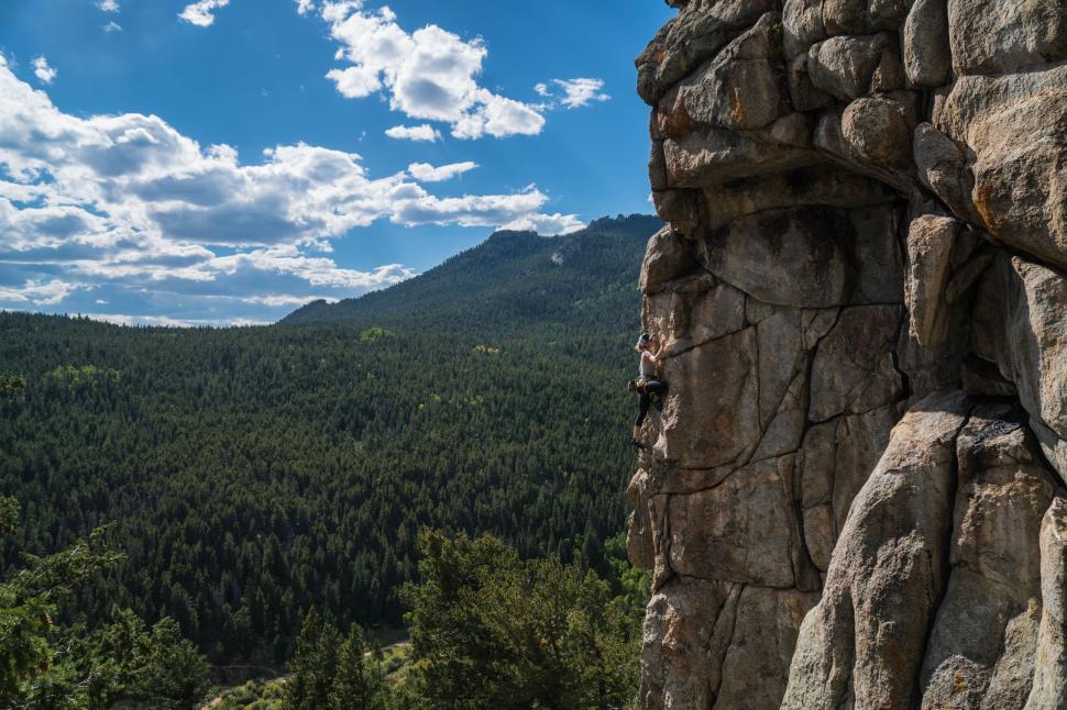 Free Image of Person climbing a steep rock face against a lush forest backdrop 