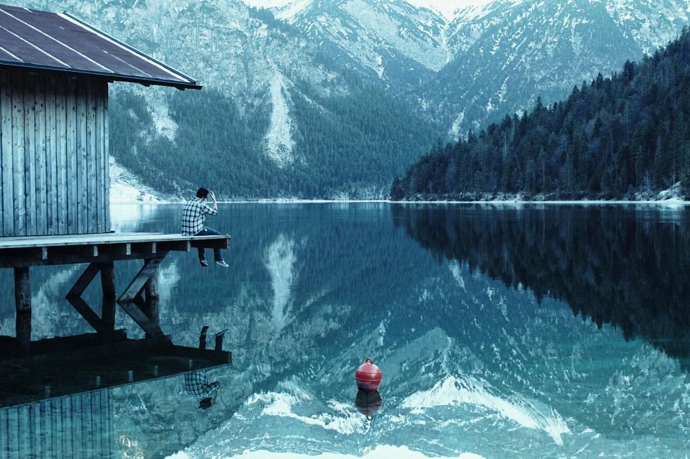 Free Image of Man sitting by the dock on a lake surrounded by mountains 