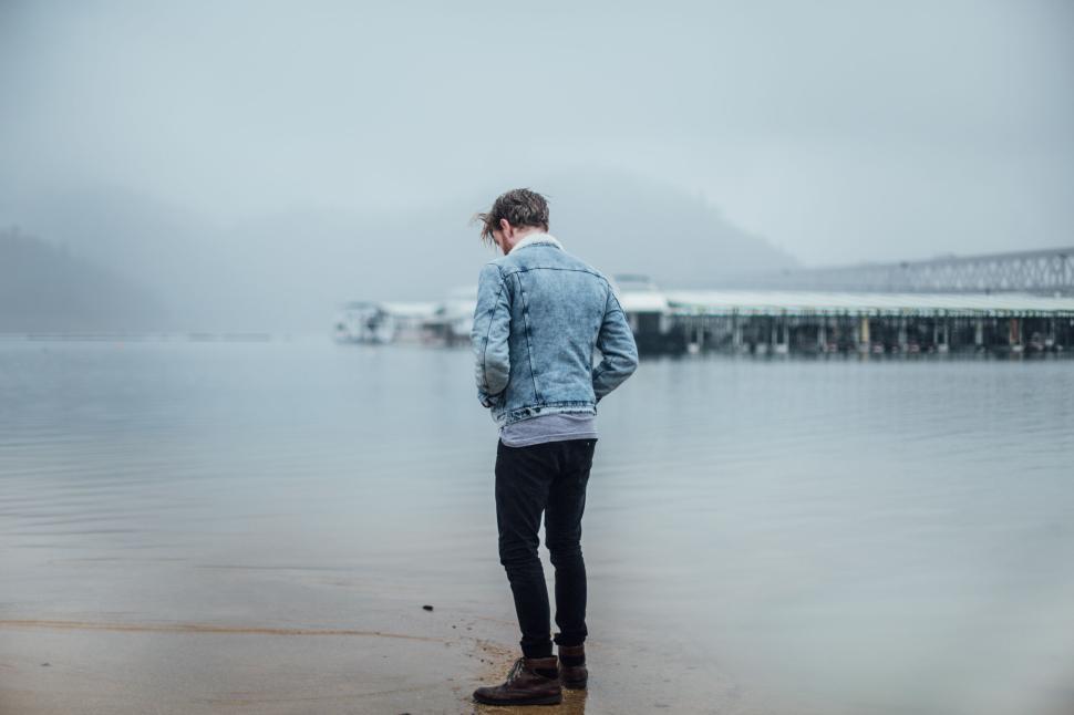 Free Image of Man standing by a lake in foggy weather wearing a denim jacket 