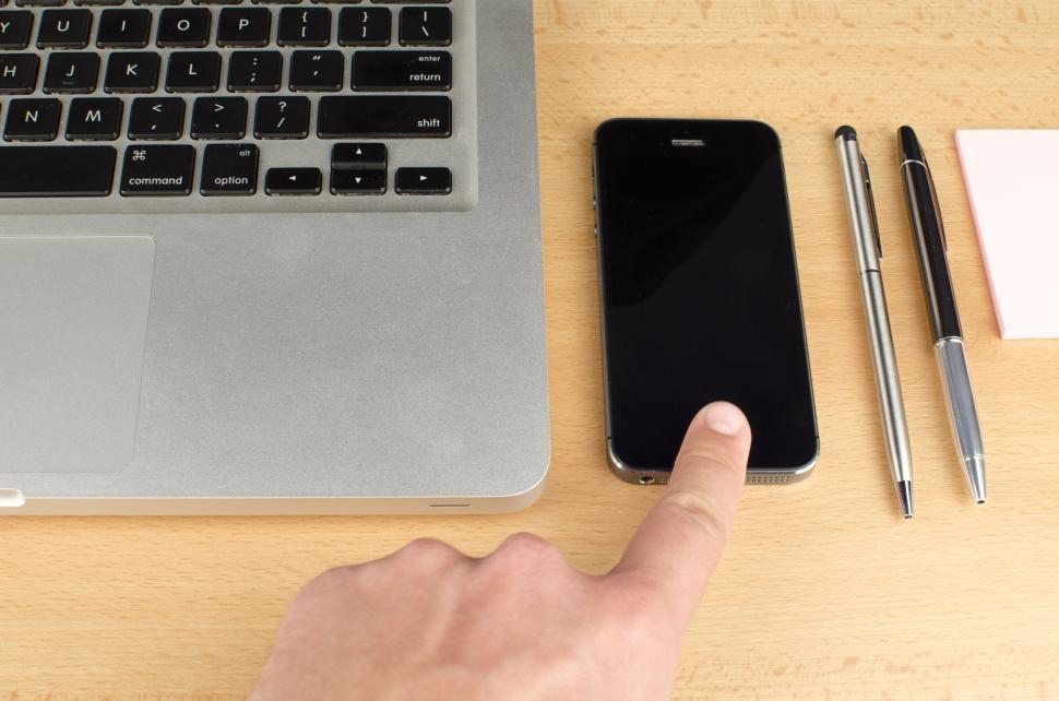 Free Image of Person touching smartphone next to laptop and pens on wooden desk 