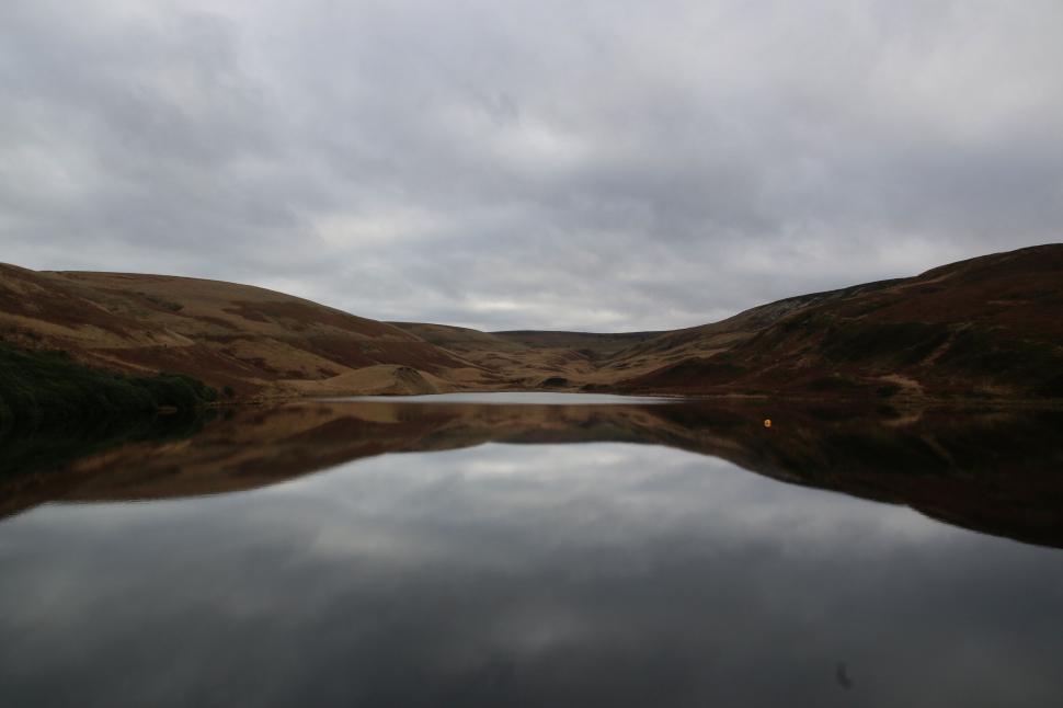 Free Image of Serene lake reflecting rolling hills under an overcast sky 