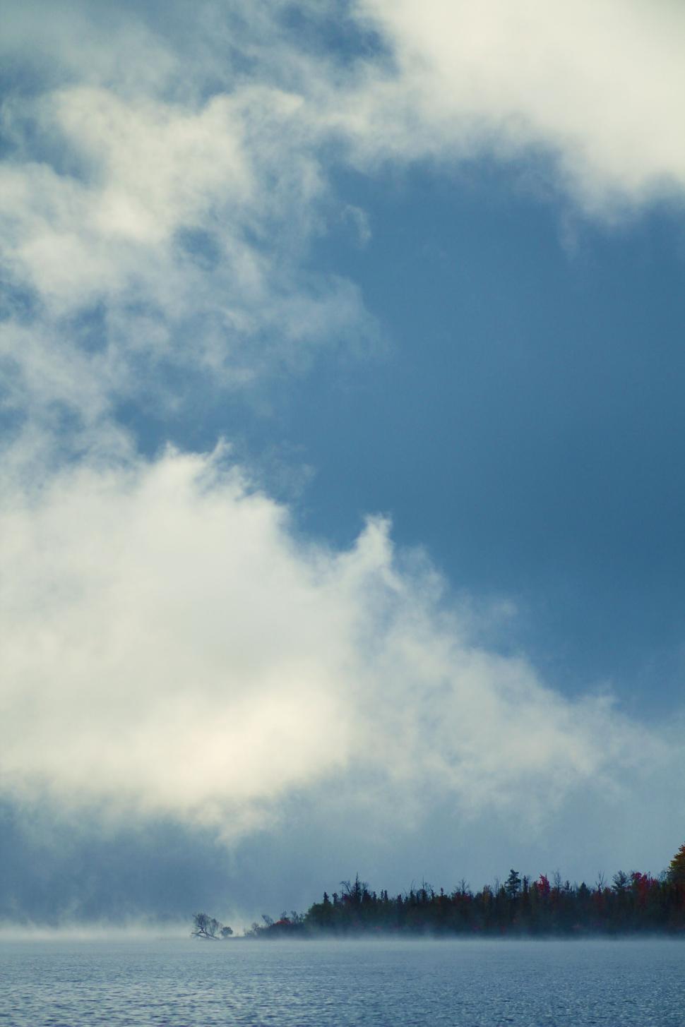 Free Image of A cloudy sky over a calm lake with distant tree line. 