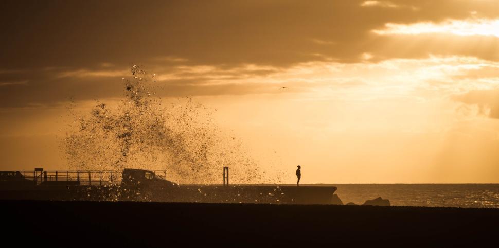 Free Image of Waves crashing along pier at sunset with person silhouetted 