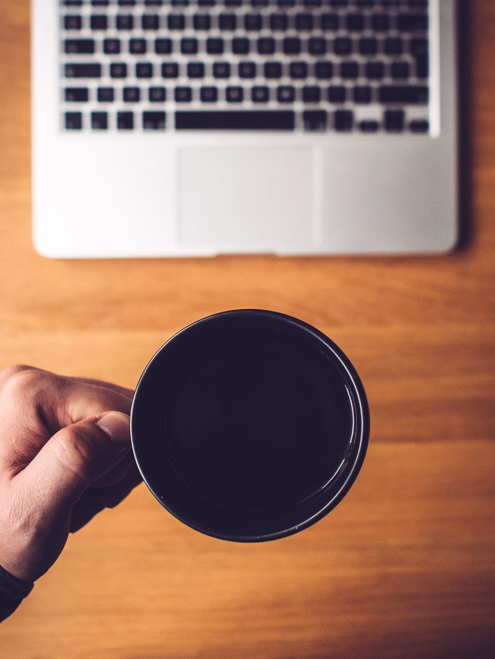 Free Image of Hand holding a coffee mug above a wooden desk surface 