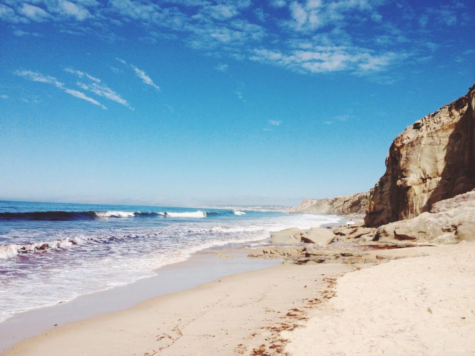 Free Image of Sunny beach with waves crashing on sandy shore, cliffs. 