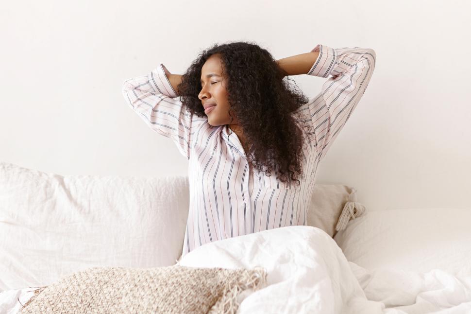 Free Image of Attractive young African American female in striped silk pajamas waking up on weekend morning, stretching body, raising hands, having joyful facial expression, sitting on bed, ready to begin new day 