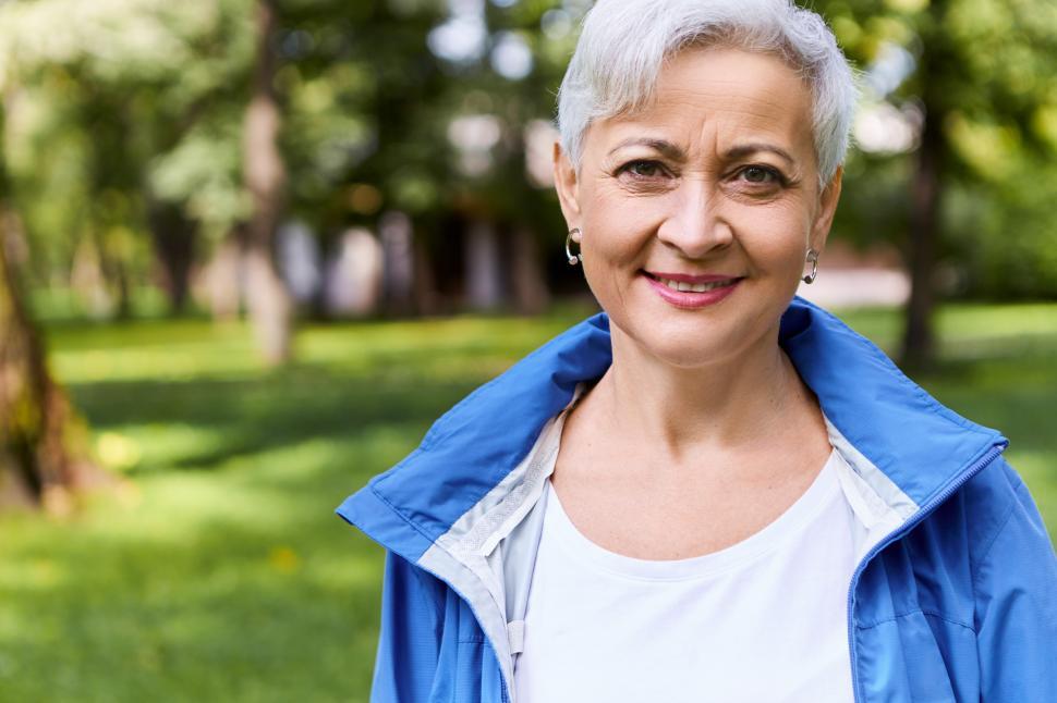 Free Image of Stylish cheerful female on retirement having nice walk in sunny forest on summer day, looking at camera with happy smile, enjoying beautiful weather and fresh air, trees and green grass in background 