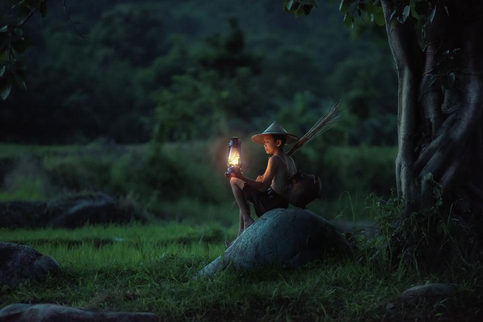 Free Image of A person in a conical hat sitting with a lantern at dusk. 
