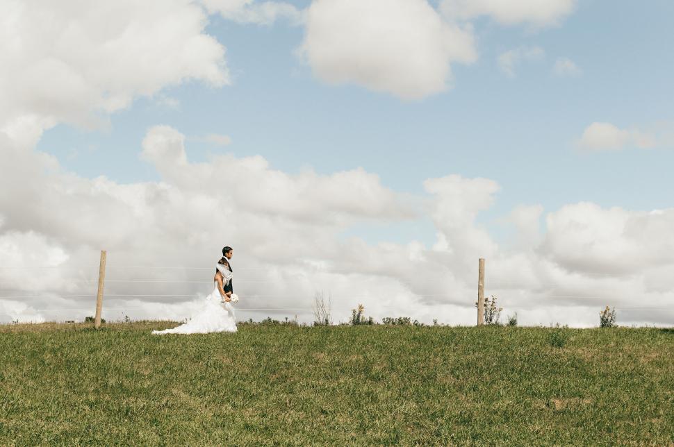 Free Image of Bride in white dress walking in open field under clouds 