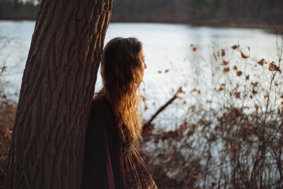 Free Image of Woman leaning on tree near peaceful lake during autumn 