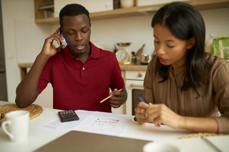 Free Image of Casually dressed dark-skinned business man sitting at kitchen table next to calculator holding pen and talking on phone with his partners, pretty young woman around him, looking at paper on desk 