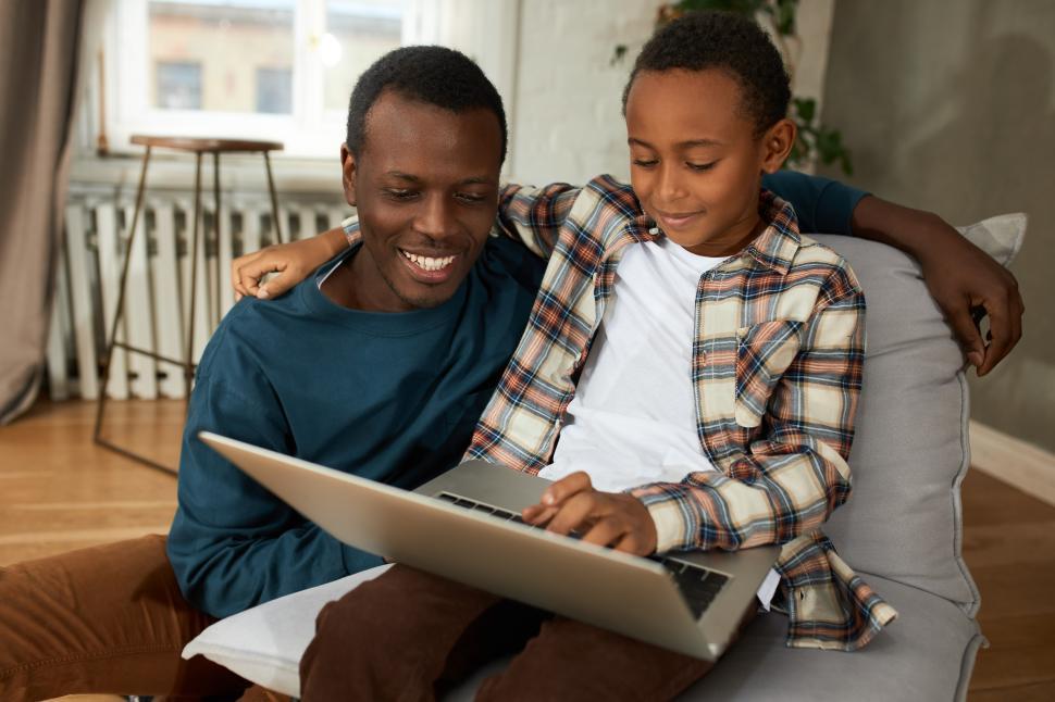 Free Image of Father and son, both dark-skinned, spending time together on Sunday watching cartoon or funny video on laptop, laughing, hugging, having fun, enjoying happy moment. Human relationships. Parenthood 