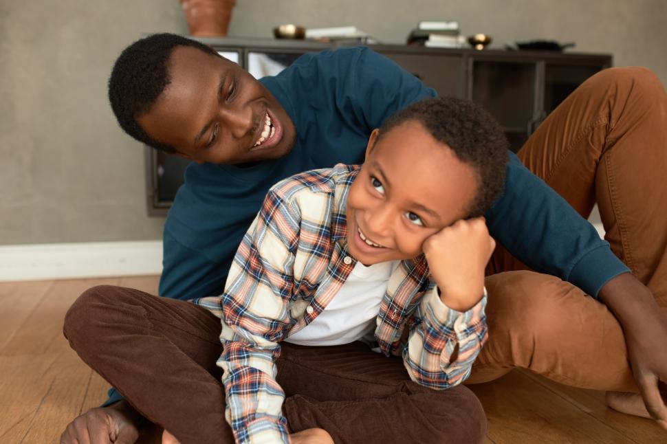 Free Image of African father cuddling his 9-years-old son while fooling around on floor in living-room against bookcase, both smiling widely, feeling happy and cheerful, enjoying moment and their warm relationships 
