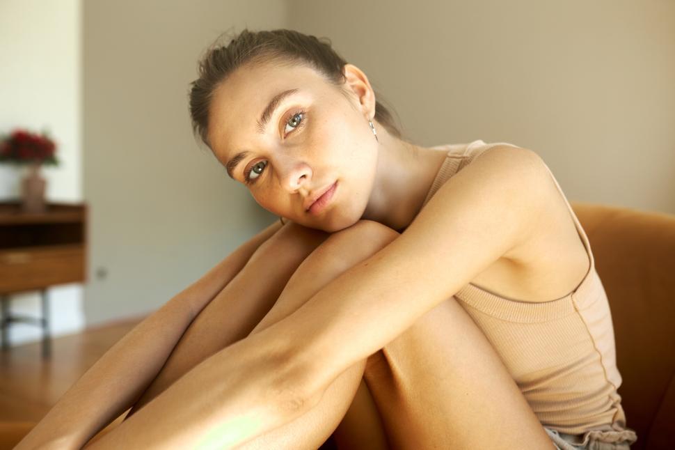Free Image of Indoor portrait of melancholic girl of 20s sitting at home feeling down and upset after splitting up with her fiance, hugging naked legs, resting chin on knees, looking at camera with sad eyes 