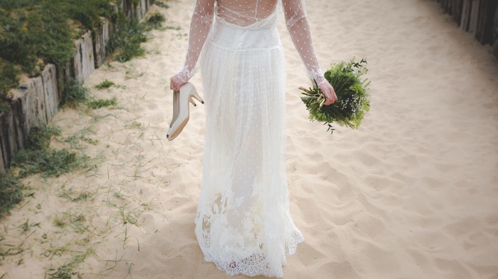Free Image of Bride holding bouquet walks barefoot on sandy beach path 