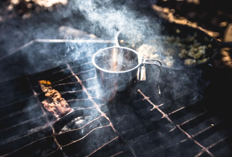 Free Image of Camping cup on a smoky grill with sizzling food nearby. 