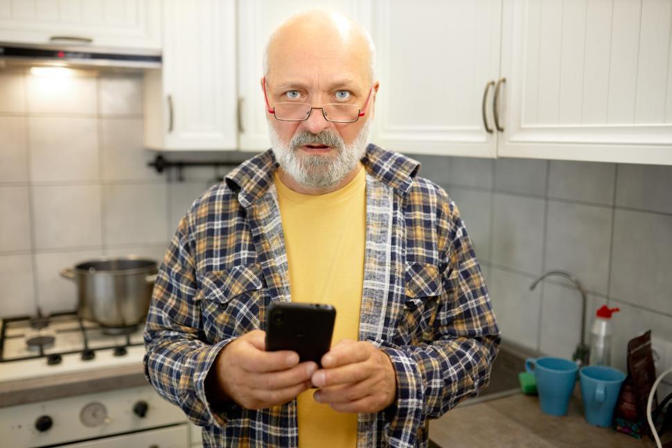 Free Image of Puzzled concerned senior male in plaid shirt standing in kitchen with smartphone in hands, trying to use mobile application to order dinner or food from supermarket, having troubles with connection 